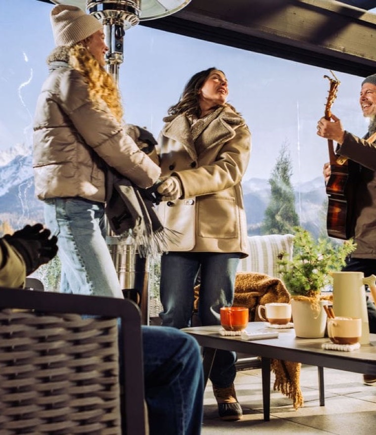 Group of friends bundled in winter coats gathering on a vinyl-enclosed patio with a snowy mountain view