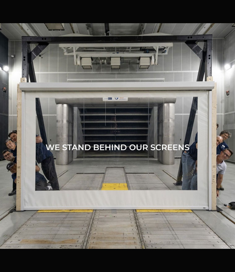 Phantom Screens team surrounding a large screen unit in a wooden test frame inside a wind tunnel testing facility, with the text We Stand Behind Our Screens overlaid